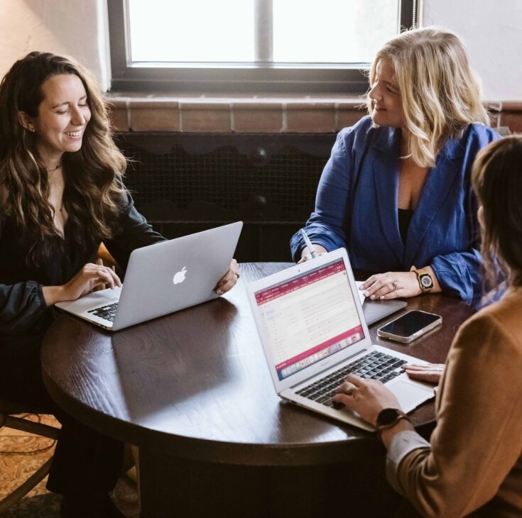 Three women are sitting at a table, working with laptops.